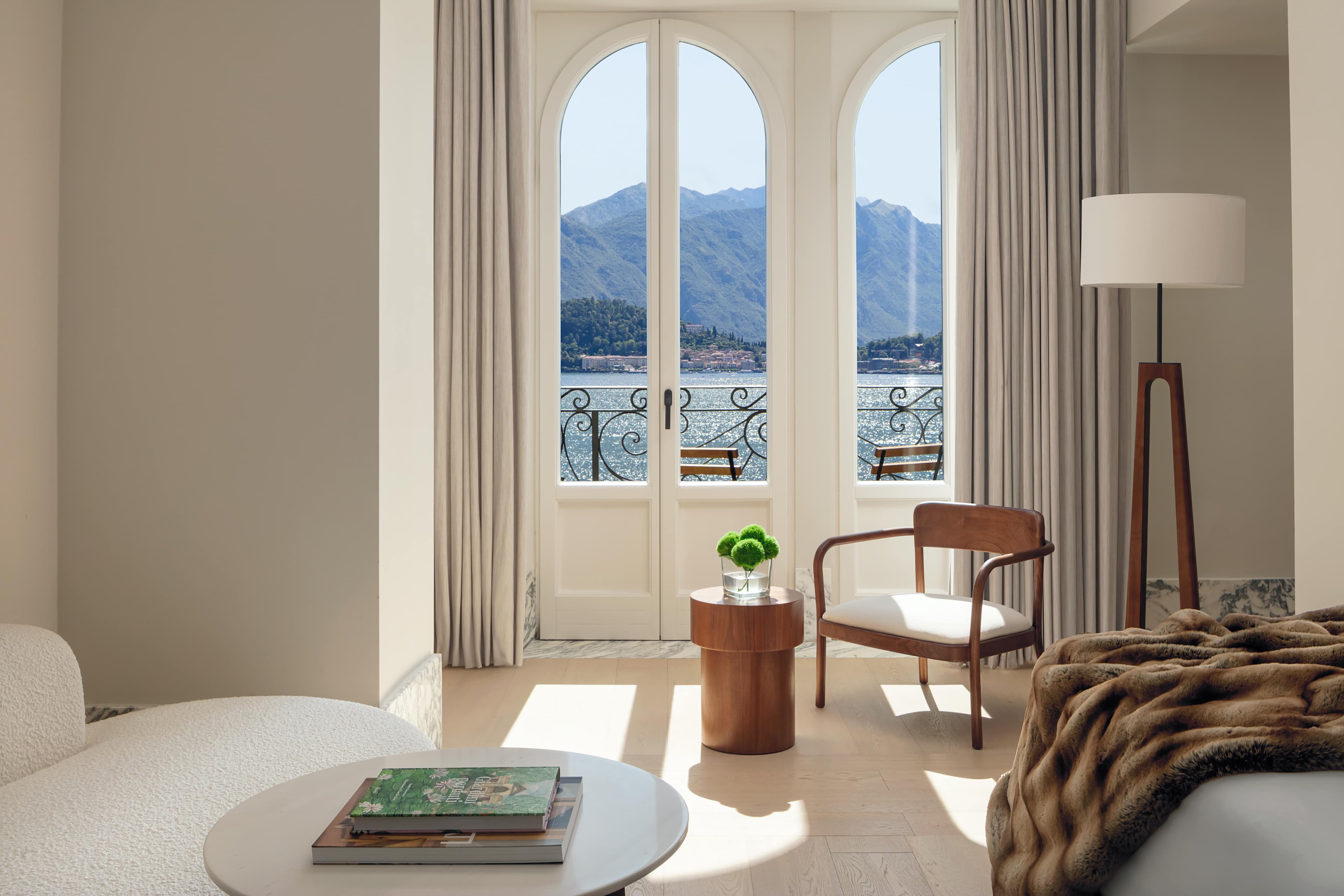 Sunlit hotel room with arched balcony doors overlooking a lake and mountains